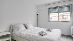 A minimalist white bedroom with a bed, bedside table, and a window with blinds. - Bright apartment with a view in Antibes (Antibes)