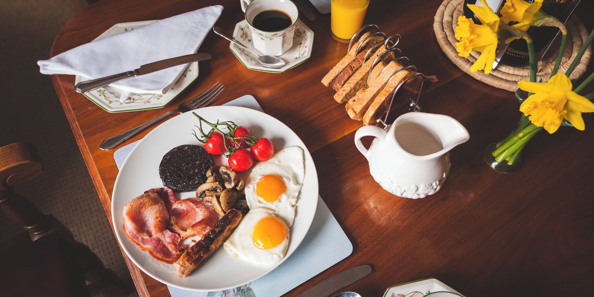A breakfast table with a plate of eggs, bacon, mushrooms, and tomatoes, a cup of coffee, a glass of orange juice, and a pitcher of milk.