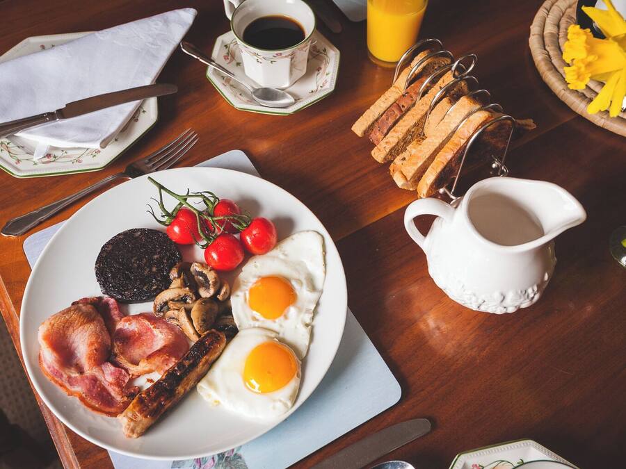 A breakfast table with a plate of eggs, bacon, mushrooms, and tomatoes, a cup of coffee, a glass of orange juice, and a pitcher of milk.