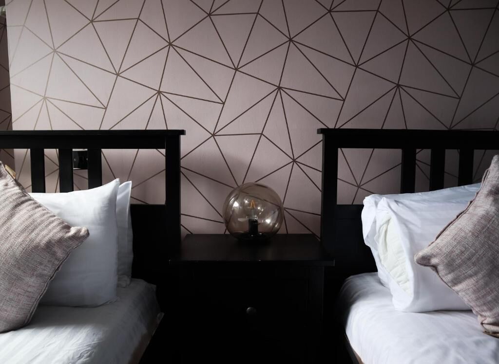 Una habitación con dos camas, una pared con diseño geométrico y una lámpara esférica en una mesita de noche.