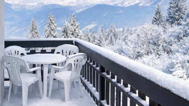 Een sneeuwbedekte veranda met een tafel en stoelen, uitkijkend op een besneeerd berglandschap.