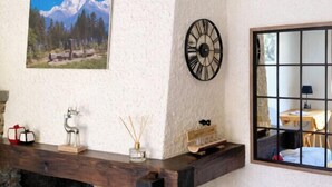 Een rustiek interieur met een stenen openhaard, een houten plank met decoratieve objecten, een wandklok en een lijst met een berglandschap.
