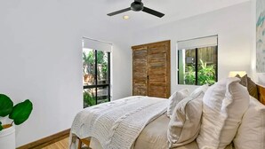 A bedroom with a bed, a ceiling fan, and two windows with views of greenery.