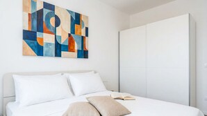 Ein ordentlich bezogenes Bett mit weißer Bettwäsche und zwei beigen Kissen, ein großes abstraktes Gemälde an der Wand und ein geschlossener Kleiderschrank.