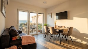 Ein modernes Wohnzimmer mit einer Couch, einem Essbereich mit Stühlen und einem Tisch, einem an der Wand montierten Flachbildfernseher und einer Glasschiebetür, die zu einer Außenpatio führt.