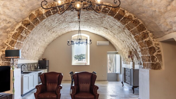 A spacious room with a vaulted stone ceiling, a chandelier, a fireplace, and a view through a window. - Casa Labisi - One Bedroom Apartment, Sleeps 4 (Noto)