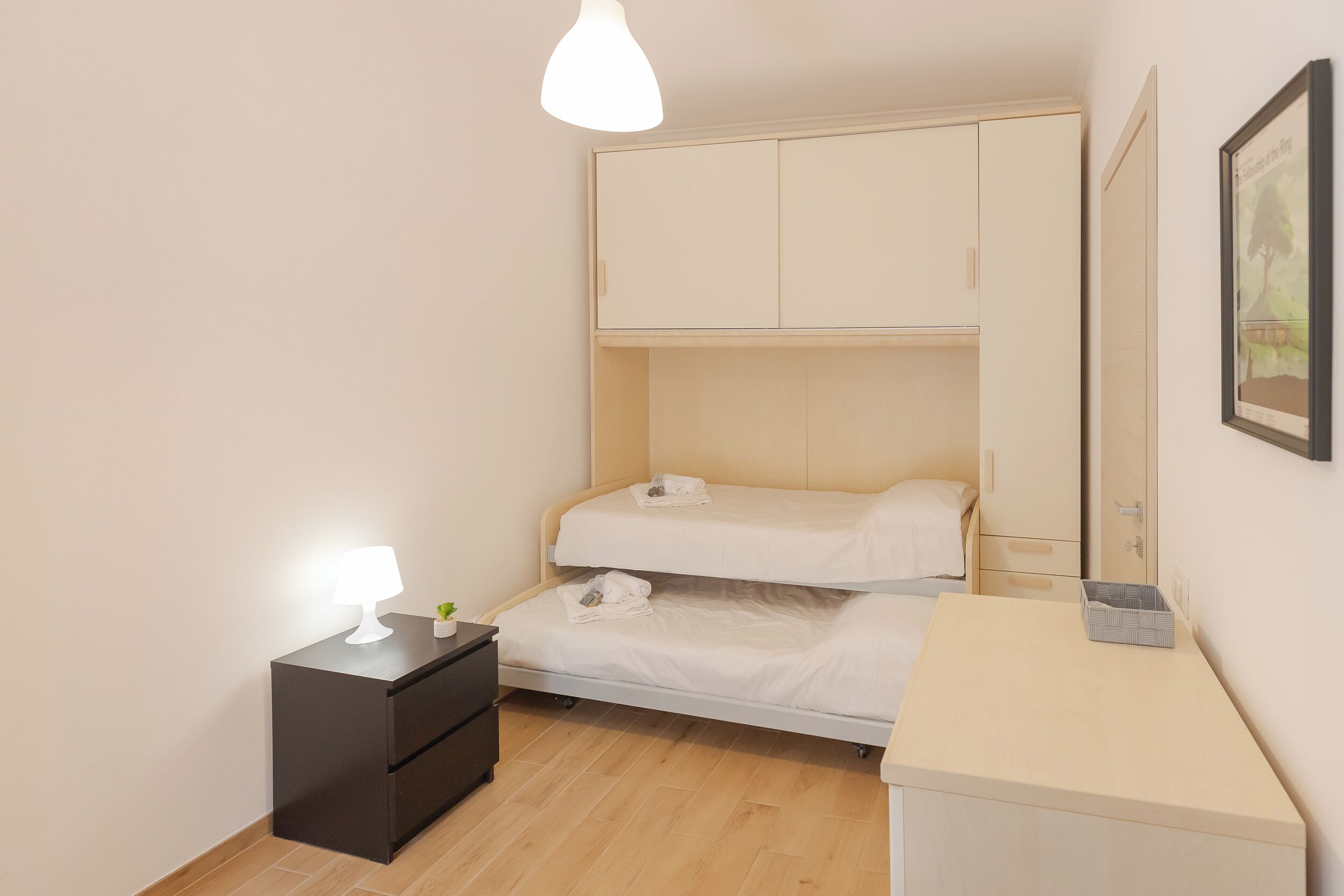 A compact bedroom with a bunk bed, a bedside table with a lamp, a small plant, and a framed picture on the wall.