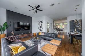 Een moderne woonkamer met een grote flatscreen televisie, een donkere bank, een salontafel en een eetgedeelte met een kroonluchter.