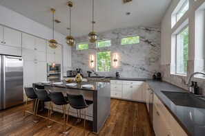 A modern kitchen with marble walls, a central island with bar stools, stainless steel appliances, and pendant lighting. - Rodeo House-Luxury-Modern-Pool-Rooftop Deck-Balcany-Skyline View-Sleeps 14 (Houston)