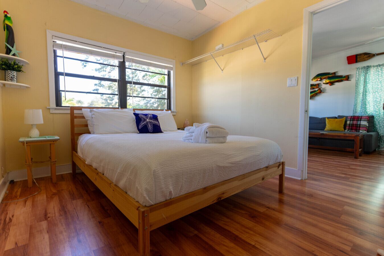 Ein Schlafzimmer mit einem hölzernen Bett, einem Fenster, einem Nachttisch und Blick auf einen Wohnbereich mit einer Couch und Vorhängen.