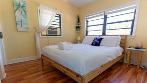 A neatly made bed with white linens and a blue pillow, a wooden nightstand with a salt lamp, and a window with white curtains. - Cindy Villa - Key Largo (Key Largo)