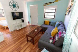 Ein Wohnzimmer mit Holzboden, einem grauen Sofa mit bunten Kissen, einem Holztisch, einem Flachbildfernseher auf einem weißen Standfuß und einem Fenster mit einem hängenden, dekorativen Fisch.