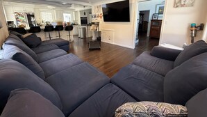 A spacious living room with a large grey sectional sofa, a flat-screen TV mounted on the wall, and a kitchen area in the background. - World Cup Stay in Easton! Elegant Suburban Escape Close to Boston Excitement (Easton)