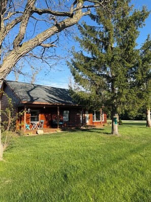 A rustic cabin with a porch, surrounded by a grassy yard and large trees. - Otsego Lake Lodge - Your next destination! (Gaylord)