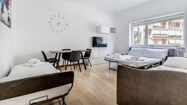 Een moderne, goed verlichte kamer met een eettafel, stoelen, een bed en een televisie.