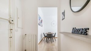 Een gang met een witte deur, een radiator, een eethoek met tafel en stoelen, en een 'WELCOME' bord op een plank.