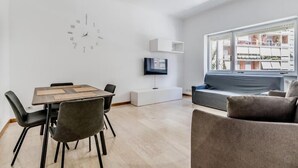 Een moderne woonkamer met een eettafel, stoelen, een bank, een televisie en een groot raam met uitzicht op gebouwen.