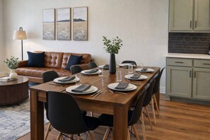 Ein Essbereich mit einem Holztisch, gedeckt für sechs Personen, umgeben von schwarzen Stühlen, einem braunen Ledersofa und einer Lampe.