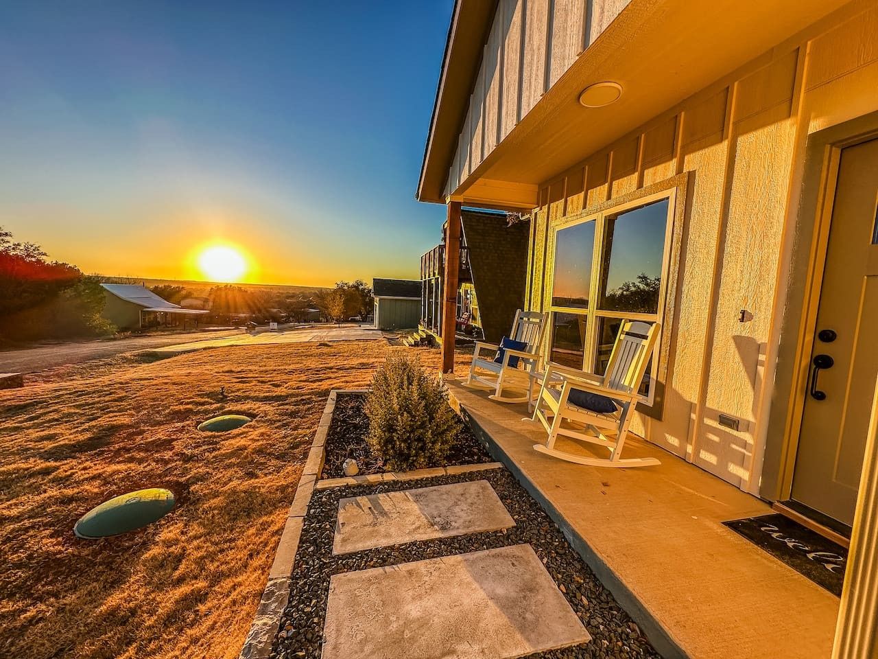 Una casa con porche cubierto, un pequeño jardín con piedras y una vista al atardecer.