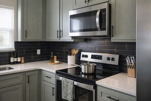 A modern kitchen with grey cabinets, a black tiled backsplash, stainless steel appliances, and a window with blinds.