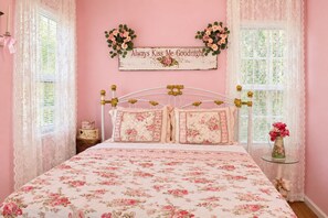 Een slaapkamer met een bloemenbedekking, een decoratief bord boven het bed en een vaas met bloemen op een nachtkastje.