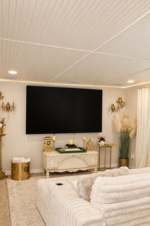 Een kamer met een grote flatscreen televisie aan de muur, een decoratieve console tafel met een klok en kaarsen, een comfortabele bank en een grote potplant.