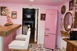 Une pièce décorée dans des tons roses, avec un vieux réfrigérateur, une table en bois, un fauteuil blanc et un miroir orné d’un cadre floral.
