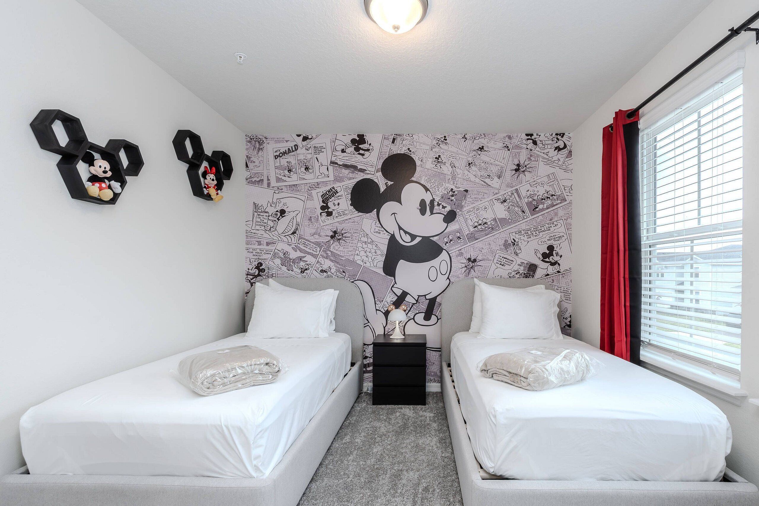 Ein Doppelzimmer mit zwei Betten, einer Wandmalerei mit Mickey Mouse und einem Fenster mit roten Vorhängen.
