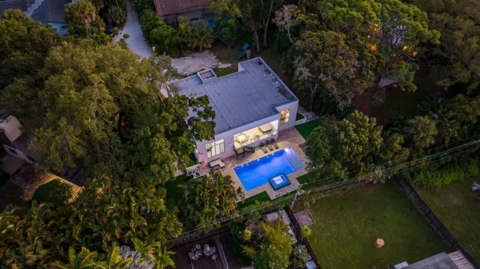 An aerial view of a modern house with a swimming pool, surrounded by dense greenery.