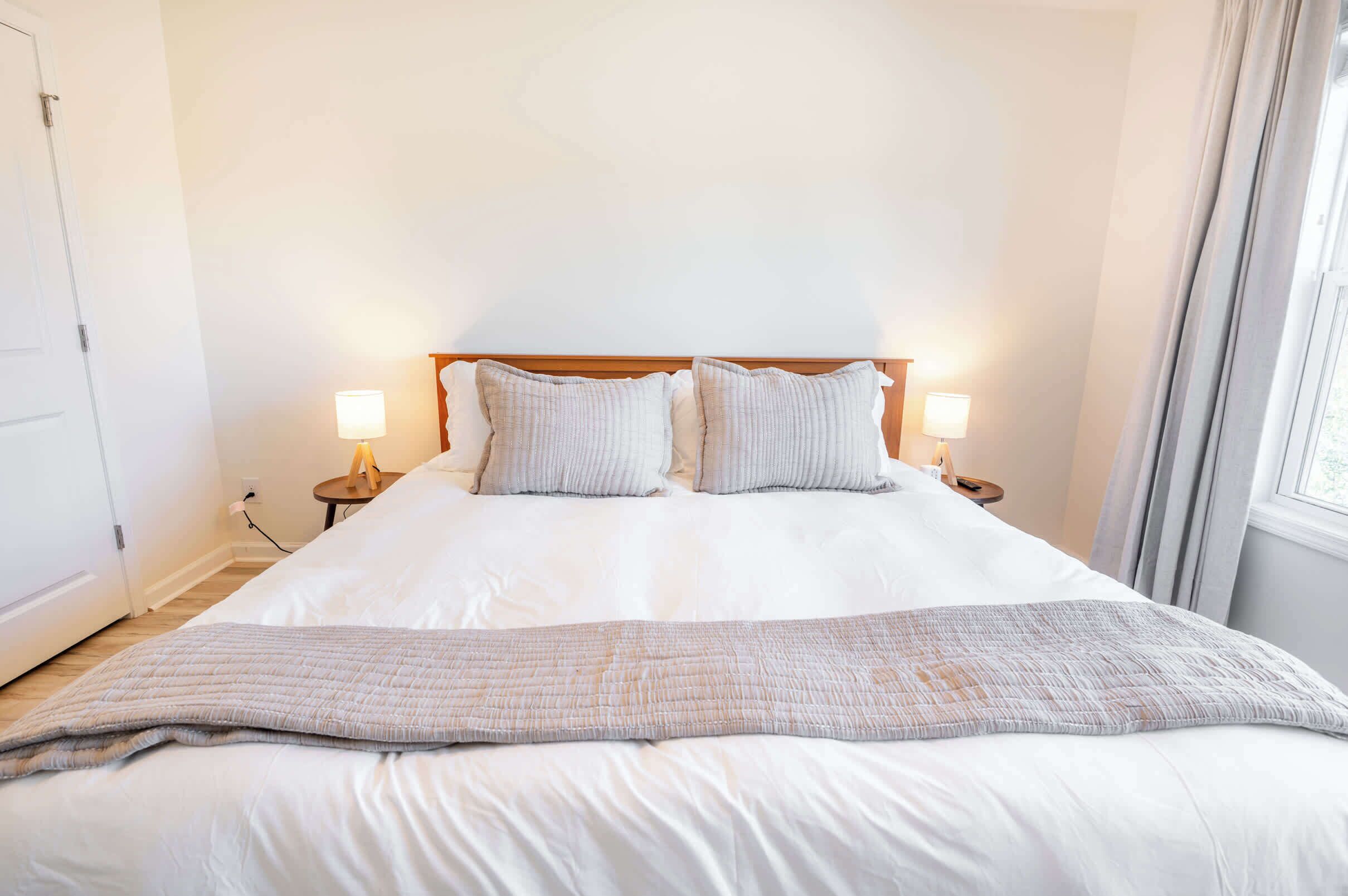 Una cama bien tendida con sábanas blancas y dos almohadas, una mesita de noche con una lámpara y una ventana con cortinas.