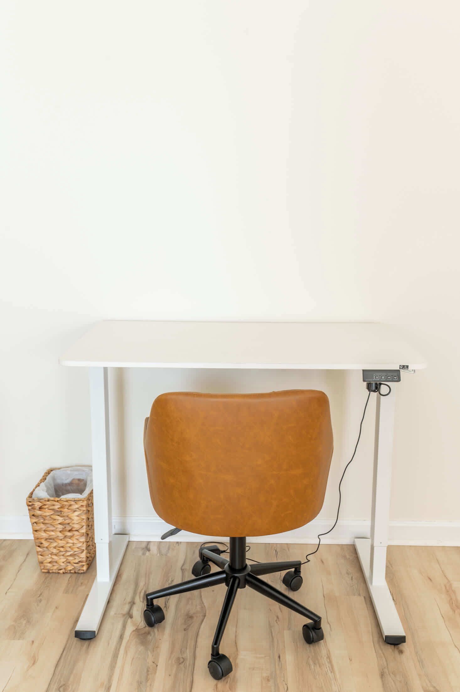 Une chaise de bureau marron, dotée d’un pied et de roulettes noirs, est placée sous un bureau blanc, avec un câble qui longe le côté du bureau.
