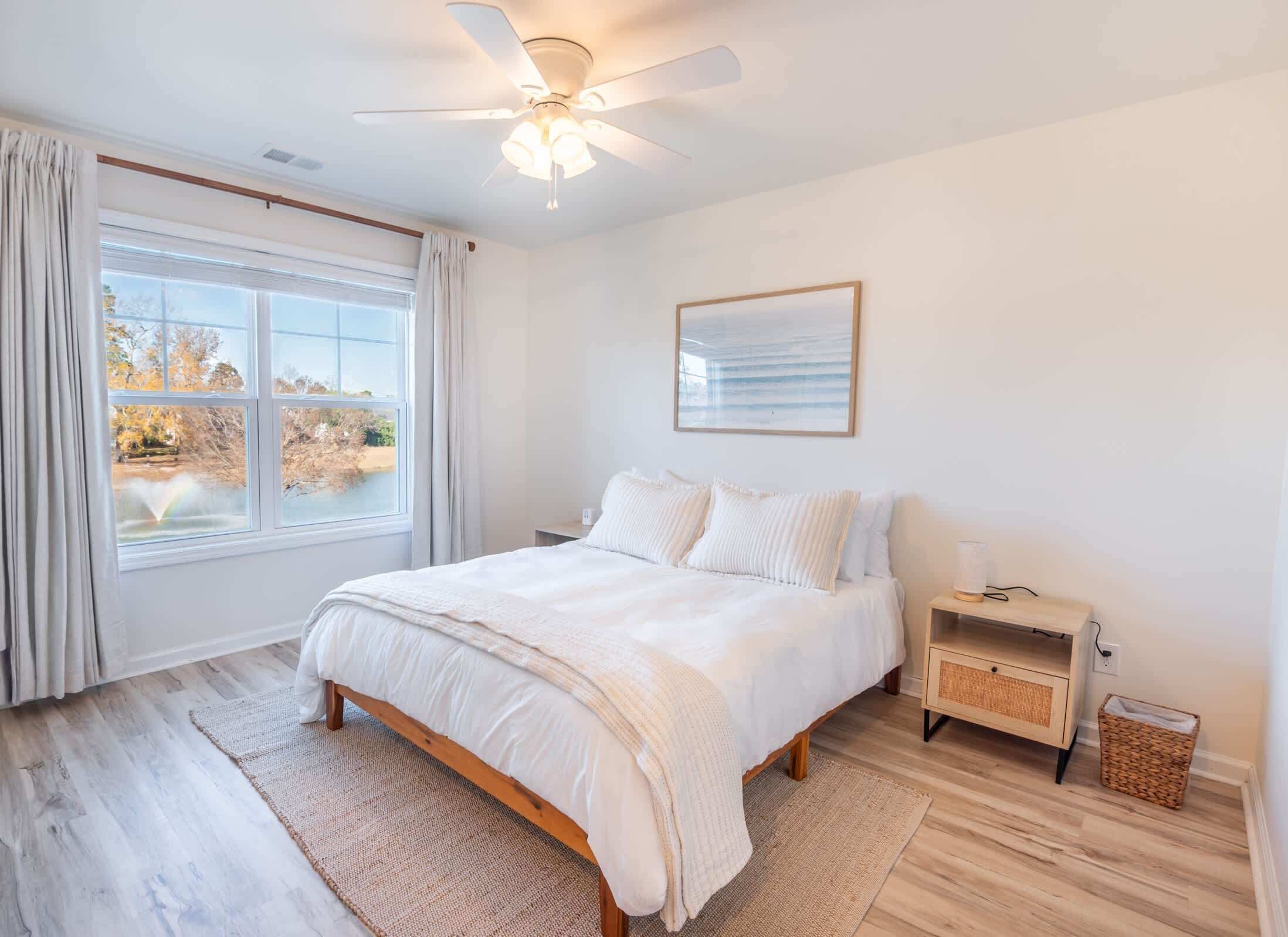 A bedroom with a bed, a nightstand, a ceiling fan, a window with curtains, and a framed picture on the wall.