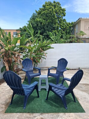 A patio with four blue plastic chairs and a small table on artificial grass, surrounded by greenery and a white wall. - Unit 2@Casa Catalina-City, Culture, & Coastline (San Juan)
