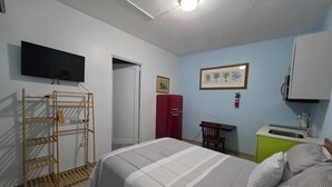 Un dormitorio con cama, armario, pequeña cocina americana, televisor montado en la pared y puerta que da a otra habitación.