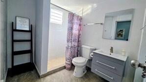 Un baño con inodoro, lavabo y tocador. Hay una ventana con persianas, una cortina de ducha y una cita enmarcada en la pared.