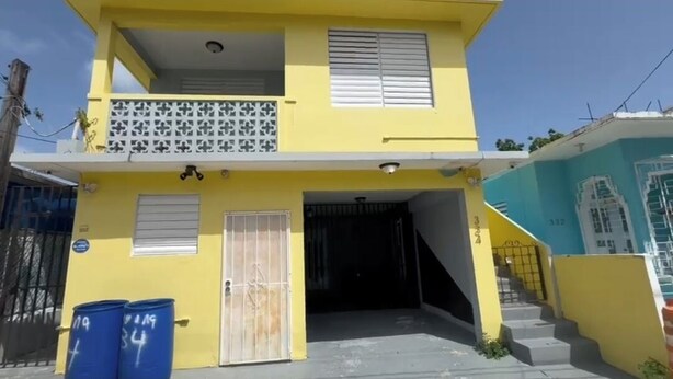 Un edificio amarillo de dos pisos con un balcón, una entrada con reja y una escalera que lleva al piso superior.