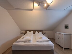 Una cama bien hecha con almohadas y toallas dobladas, una mesita de noche con lámpara, un aparador y un candelabro en el techo.