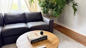 Ein Wohnzimmer mit einer schwarzen Ledersofa, einem runden Holztisch mit einem Buch und einer Fernbedienung, sowie einer großen Grünpflanze.