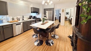 Eine moderne Küche mit Edelstahlgeräten, ein Essbereich mit einem weißen Tisch und vier Stühlen sowie ein Gang, der zu einem Schlafzimmer führt.