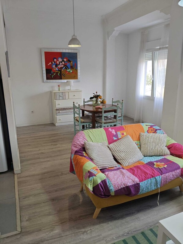 Un dormitorio con una colcha de patchwork colorida, un tocador blanco, un cuadro en la pared y una ventana con cortinas traslúcidas.