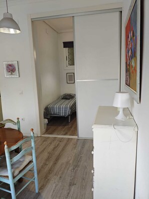 Una habitación pequeña y funcional con cama, escritorio y silla. Cuenta con una ventana con persiana, un cuadro en la pared y una lámpara sobre el escritorio.