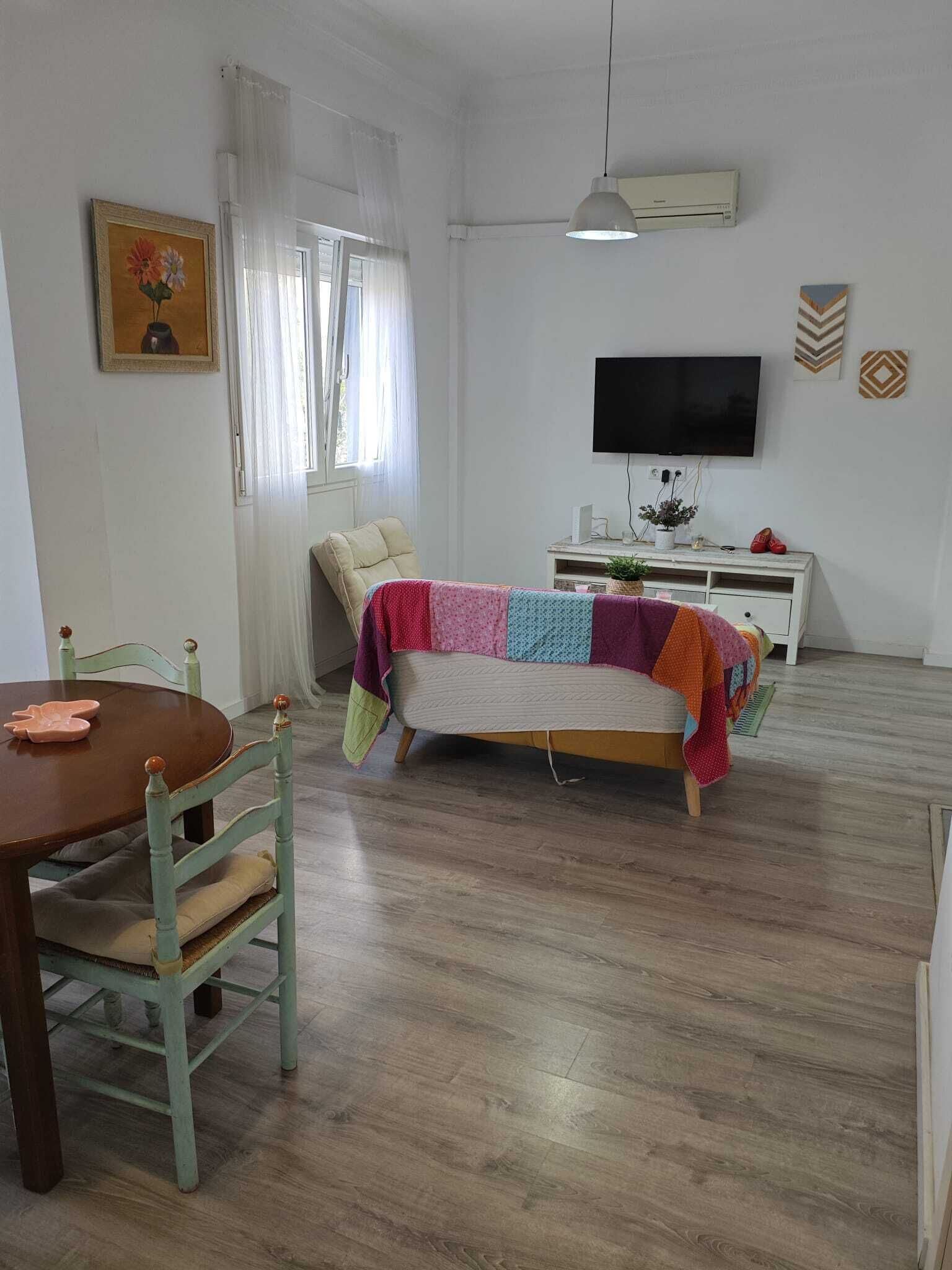 Una habitación con piso de madera, una cama con colcha colorida, un televisor en la pared, una mesa de comedor con sillas y una ventana con cortinas.