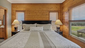 Ett sovrum med en sänggavel i trä, två sänglampor, en stor säng med mönstrat sänglinne och utsikt över en gräsyta genom ett fönster.