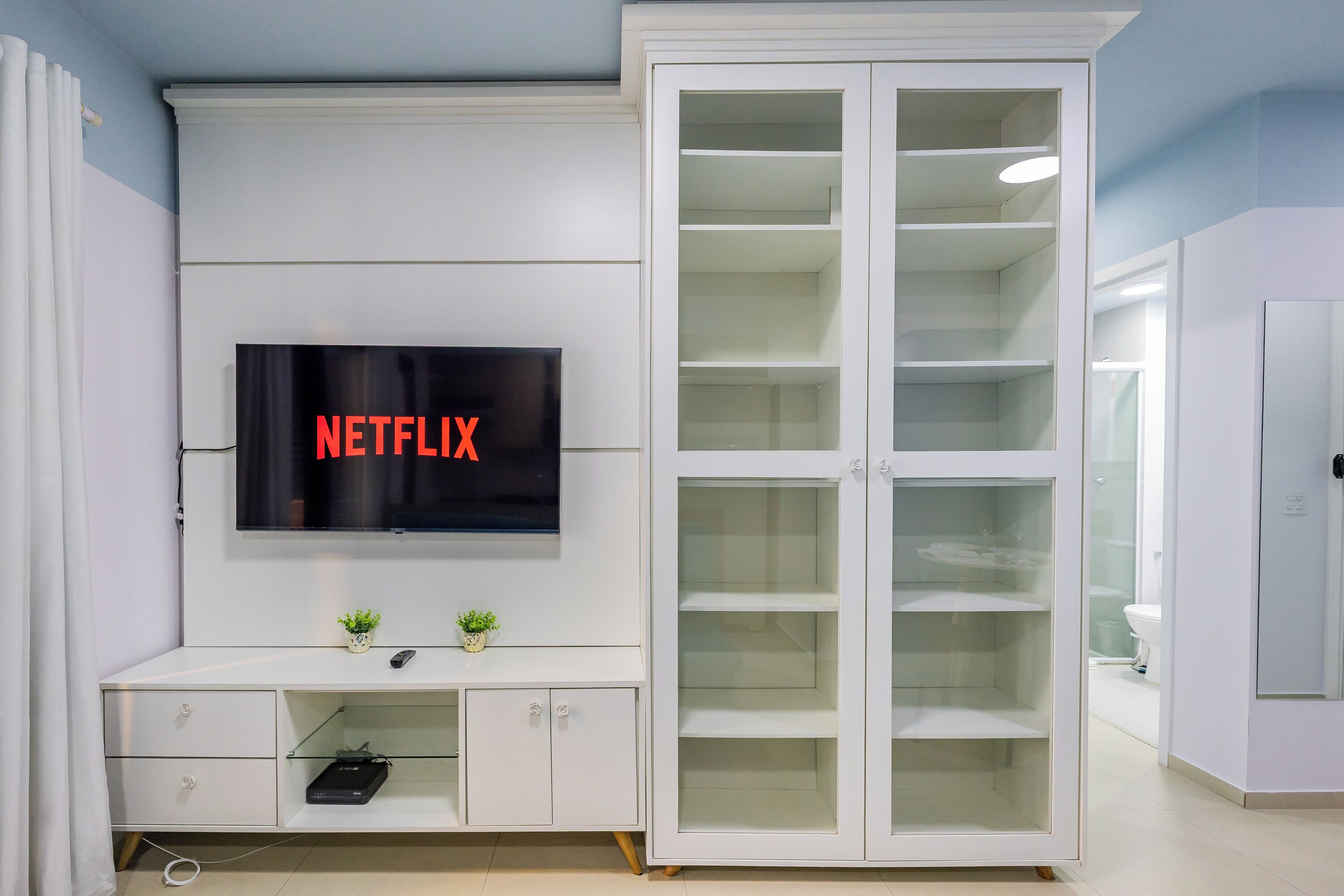 Ein modernes Wohnzimmer mit an der Wand montiertem Fernseher, einem weißen Schrank mit Glastüren und einer kleinen Pflanze auf dem Schrank.