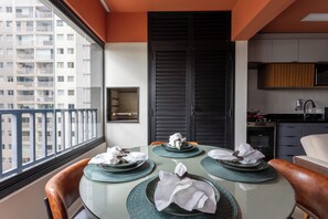 Une table à manger dressée avec des assiettes et des serviettes, un coin cuisine avec des placards et des appareils électroménagers, et une vue sur un immeuble par la fenêtre.