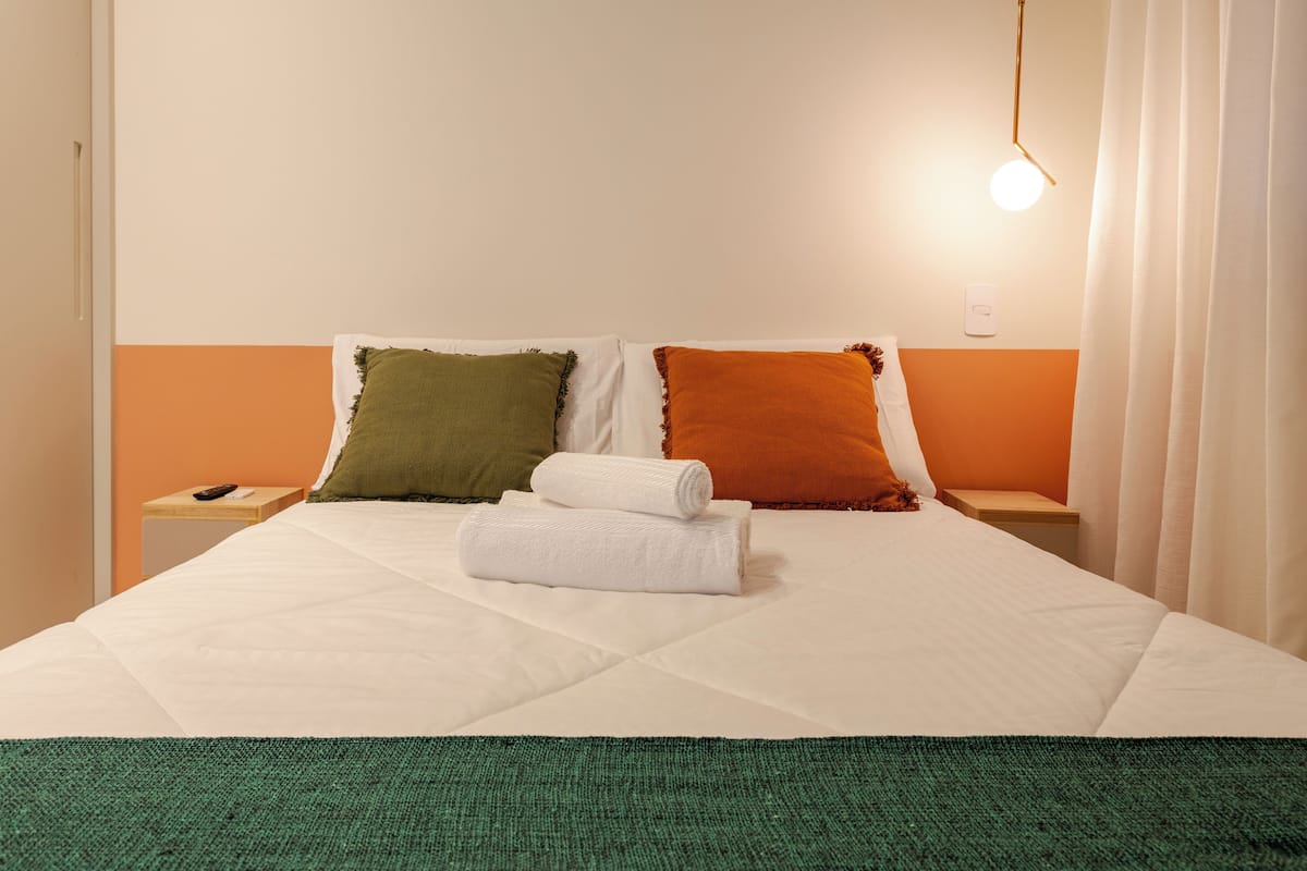 Un lit bien fait, avec une couette blanche, deux oreillers verts et orange, et une serviette blanche roulée posée sur le lit.