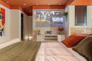 Un dormitorio con una cama, un televisor montado en la pared, una mesita de noche blanca y una ventana con persianas.