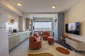 Ein modernes Wohnzimmer mit Sofa, Hocker und Fernseher, eine Küche mit weißen Schränken und ein großes Fenster mit Blick auf die Stadt.