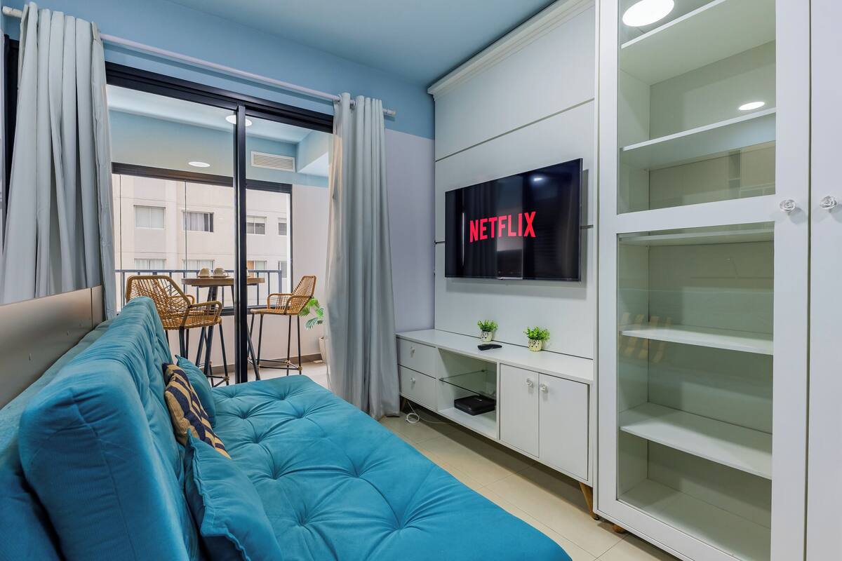 Un salon moderne avec un canapé bleu, un téléviseur fixé au mur et un balcon offrant une vue sur les immeubles.