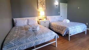 A room with two beds, a nightstand with a lamp, a wall-mounted light, and a door.